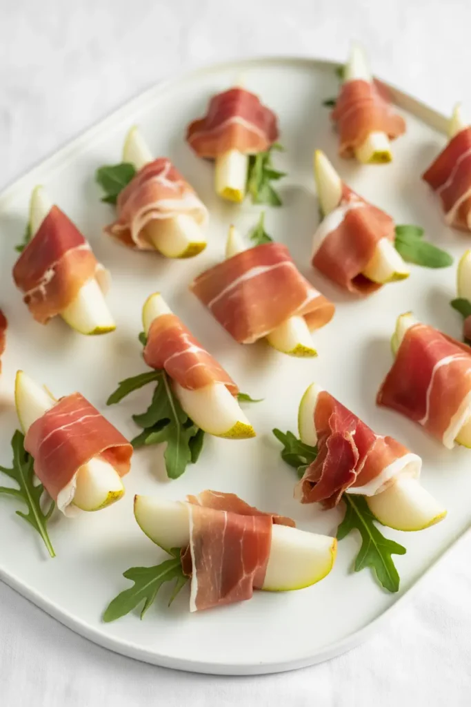 Fresh pear wedges and peppery arugula wrapped in thin slices of salty prosciutto, arranged as a no-cook appetizer on a white platter.