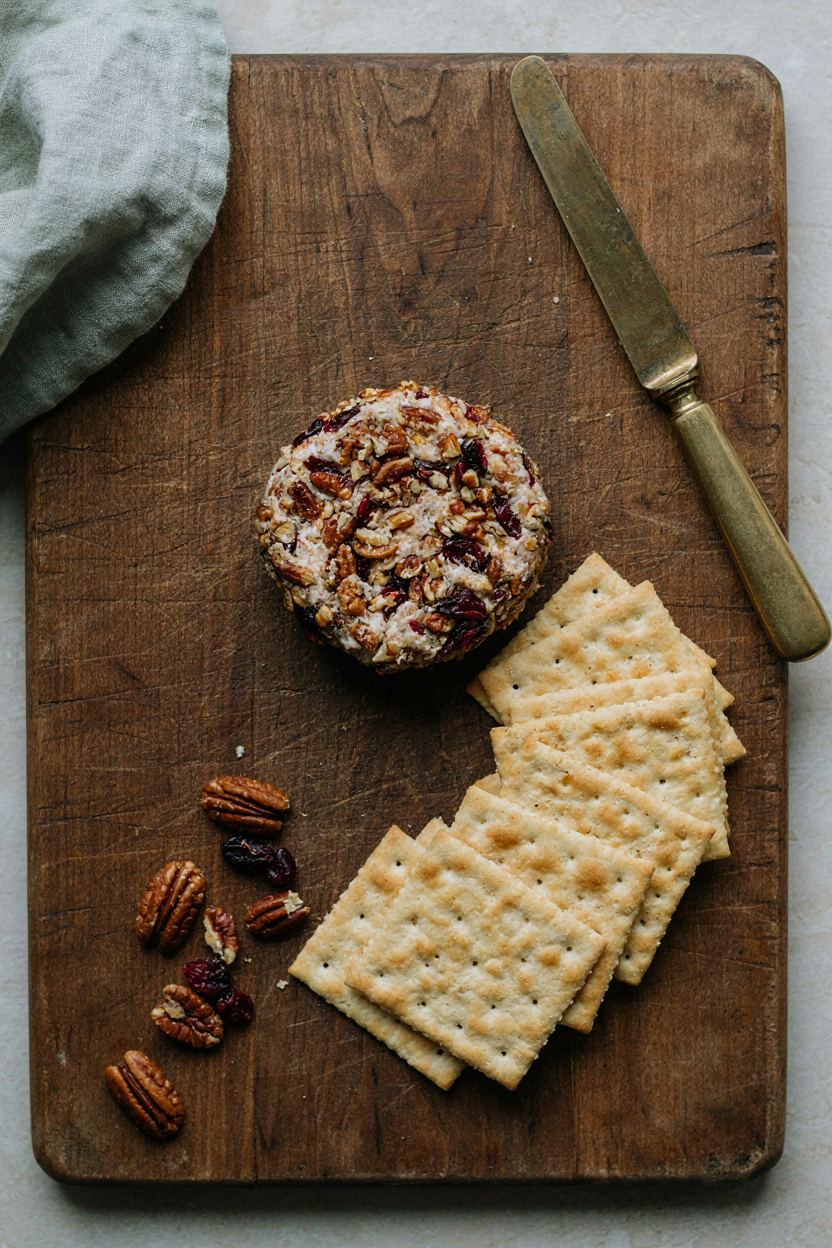 Cheese ball coated in chopped pecans and dried cranberries on a rustic board with crackers fanned around