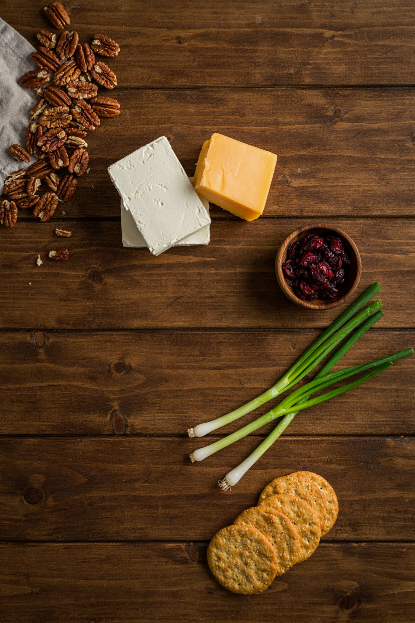 Cheese ball ingredients on rustic table: block cream cheese, sharp cheddar, pecans, cranberries, green onions, crackers