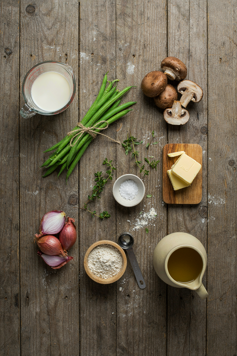 Green bean casserole ingredients on rustic table: green beans, mushrooms, shallots, butter, flour, half-and-half